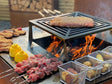 byJEMA Feuerstelle aus Cortenstahl mit sichtbaren Flammen – Fleisch, Grillspieße und Kartoffeln über offenem Feuer