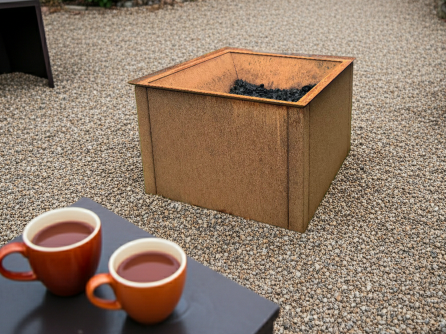 Quadratische Feuerstelle aus Cortenstahl auf Kiesfläche im Garten, mit Asche und Glut, geeignet für Terrasse und Außenbereich