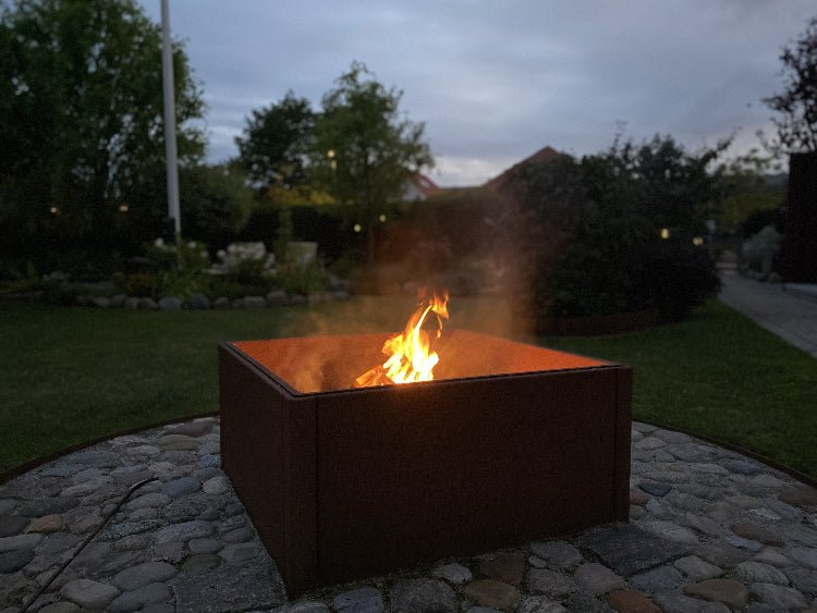 Quadratische Feuerschale aus Cortenstahl mit offener Flamme in einem Garten bei Abenddämmerung, platziert auf einer runden Fläche aus Natursteinen.