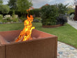 Quadratische Feuerschale aus Cortenstahl mit lebendigem Feuer, platziert in einem gepflegten Garten mit Rasen und Pflanzbeeten.